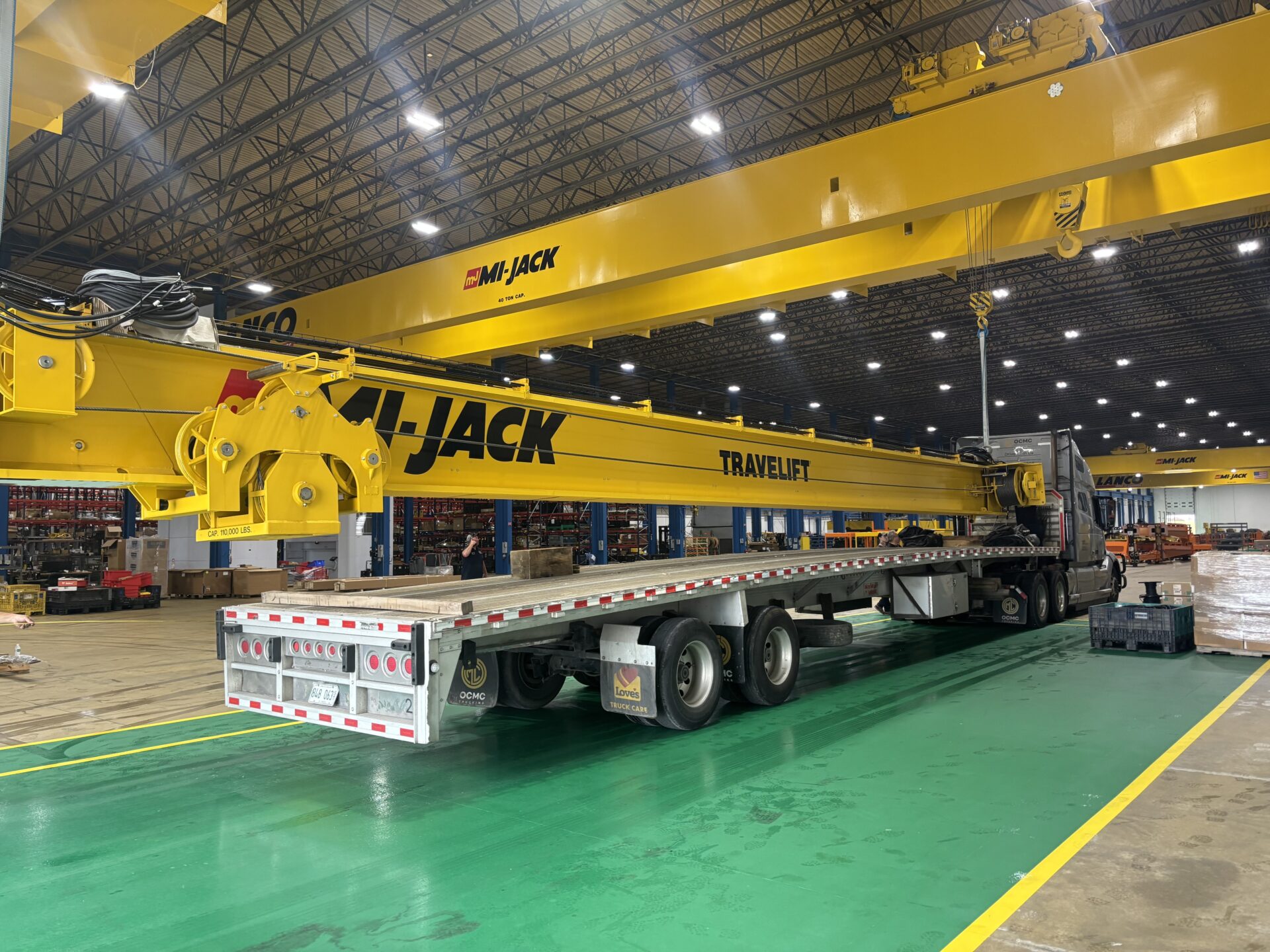 A yellow Mi-Jack Travelift rubber-tired gantry crane being loaded onto a flatbed truck inside a Mi-Jack manufacturing facility.